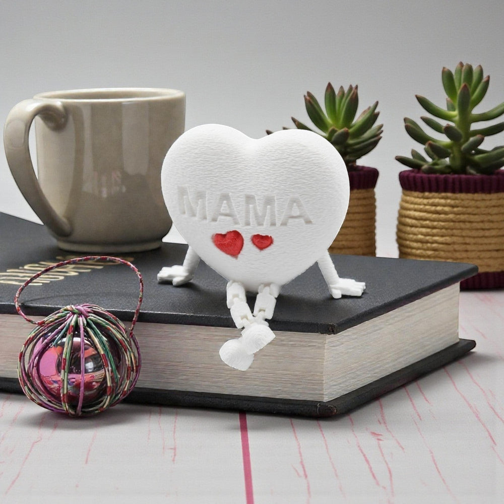 Tom Goh white heart figurine with "MAMA" embossed and two red hearts, articulated legs, sitting on a book.