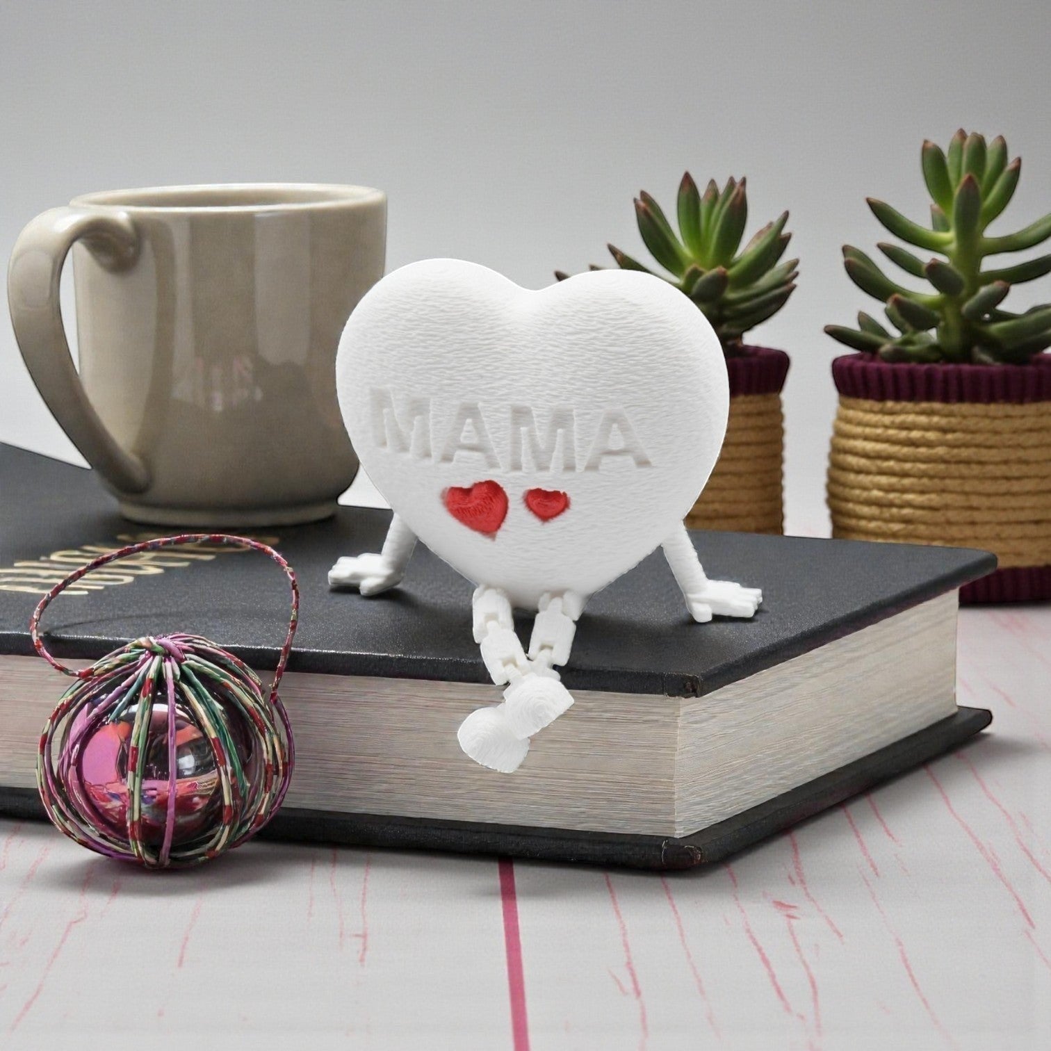 Tom Goh white heart figurine with "MAMA" embossed and two red hearts, articulated legs, sitting on a book.
